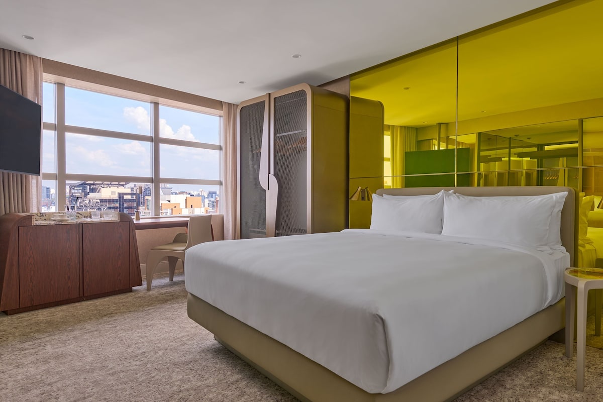 A hotel room with a large bed, a desk, a chair, a TV, and a city view.