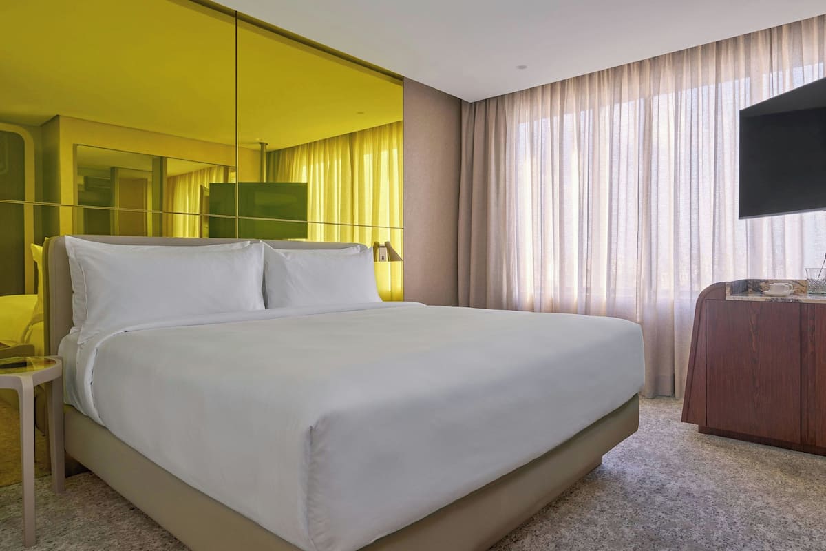 A hotel room with a large bed, a bedside table, a wooden dresser, a flat-screen TV, and a large window with curtains.