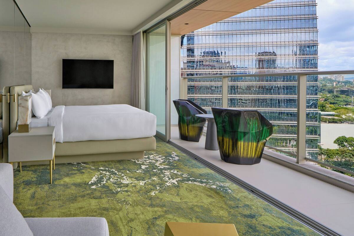 A modern hotel room with a bed, a TV, a balcony with a view of the city, and a green patterned rug.