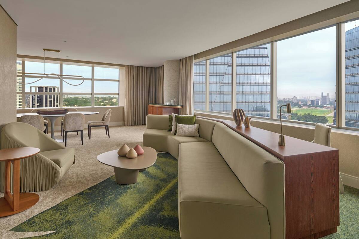 A modern hotel room with a large window, a sofa, a dining area, and a small kitchen.