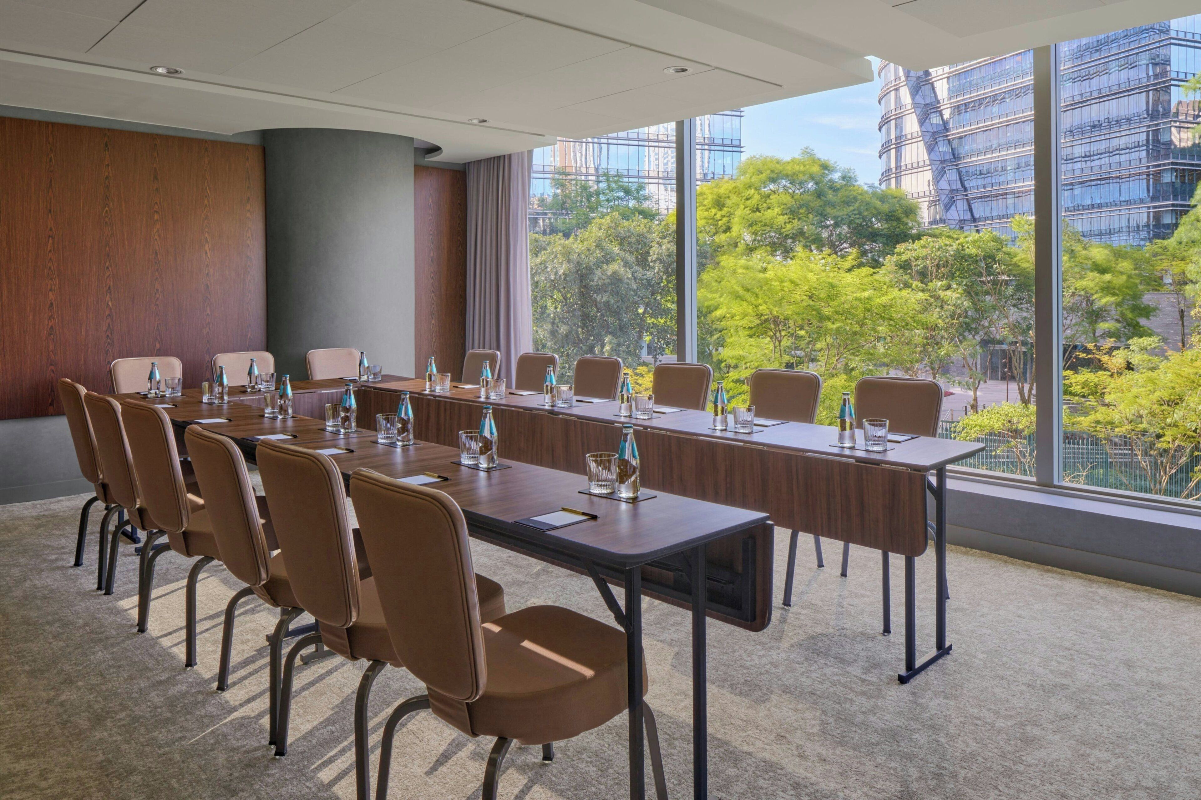 Un salón de conferencias con una larga mesa, sillas marrones y amplios ventanales que ofrecen vistas a edificios y árboles.