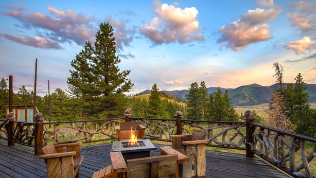 A wooden deck with a fire pit, benches, and a rustic railing overlooking a mountainous landscape.