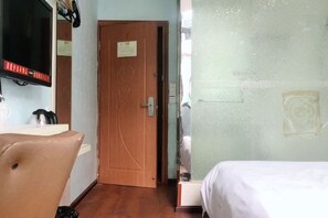 Quarto de hotel com porta de madeira, uma cama, uma cadeira e uma televisão na parede.