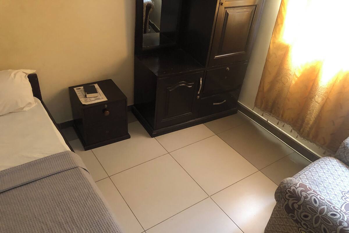 A hotel room with a bed, a small desk with a telephone, a dark wooden dresser, and a patterned armchair.