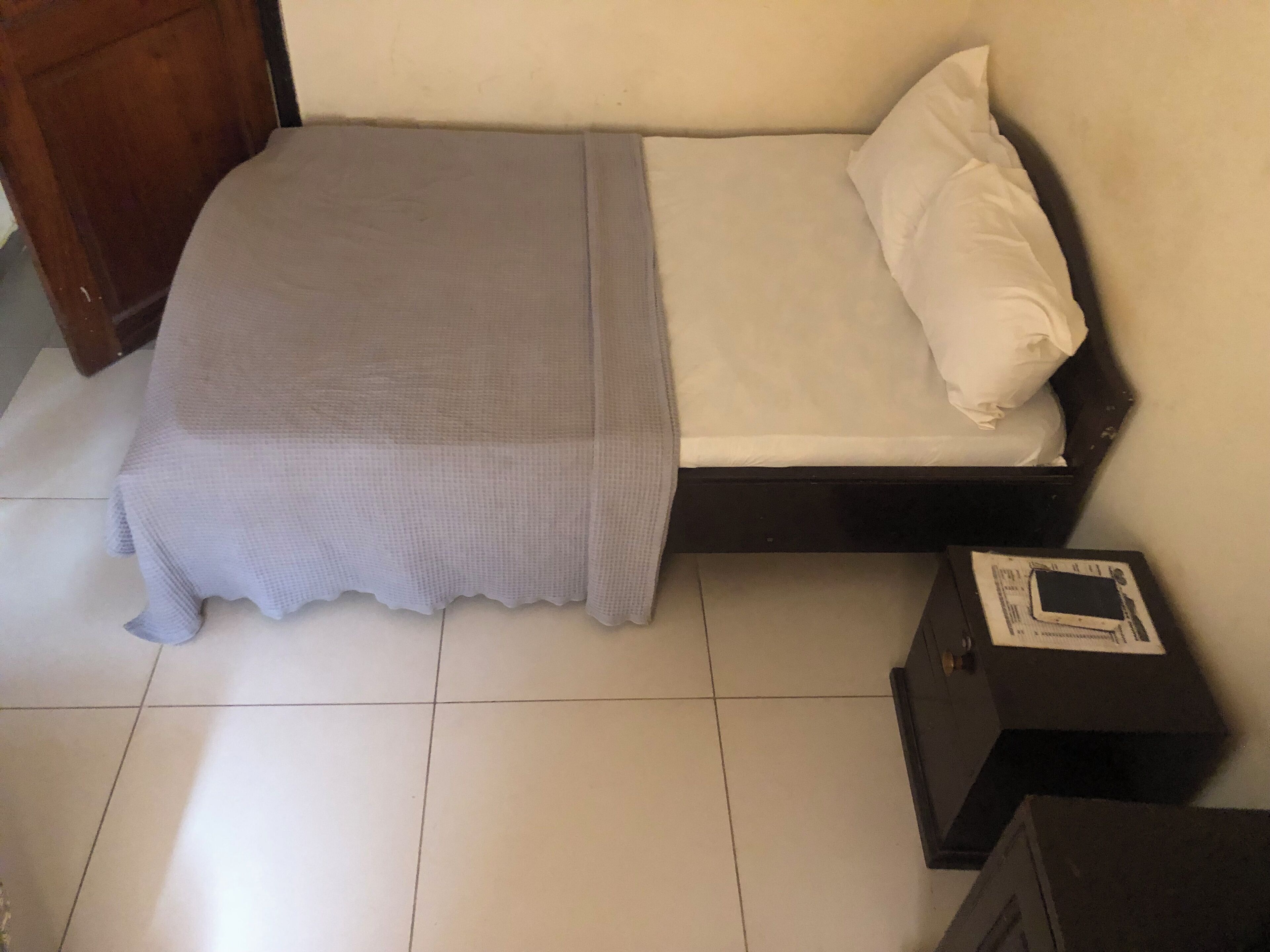 Une petite chambre avec un lit simple, une couvre-lit grise, des oreillers blancs et une tête de lit en bois. Une petite table avec une boîte dessus est visible sur la droite.