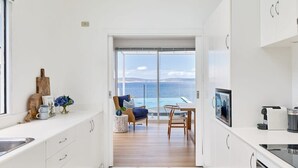 A modern kitchen with white cabinetry, a dining area with a view of the sea, and a small seating area with a table and chairs. - Waterfront Oasis with Infinity Pool and Stunning River Views (Taroona)