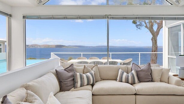 A modern living room with a large window overlooking a scenic view of the sea and hills. - Waterfront Oasis with Infinity Pool and Stunning River Views (Taroona)