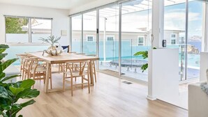 A spacious room with a wooden dining table and chairs, large sliding glass doors leading to a pool area, and a view of the surrounding buildings. - Waterfront Oasis with Infinity Pool and Stunning River Views (Taroona)