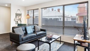 Un salon moderne avec un canapé en cuir noir, une table basse en verre et une vue dégagée sur l’extérieur grâce à de grandes fenêtres.