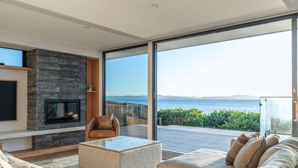 Un salon moderne avec une cheminée en pierre, un grand canapé, une table basse et une vue sur la mer grâce à de grandes portes vitrées.
