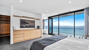 Une chambre moderne avec un grand lit, une commode en bois et une vue sur l’océan grâce à de grandes portes vitrées.
