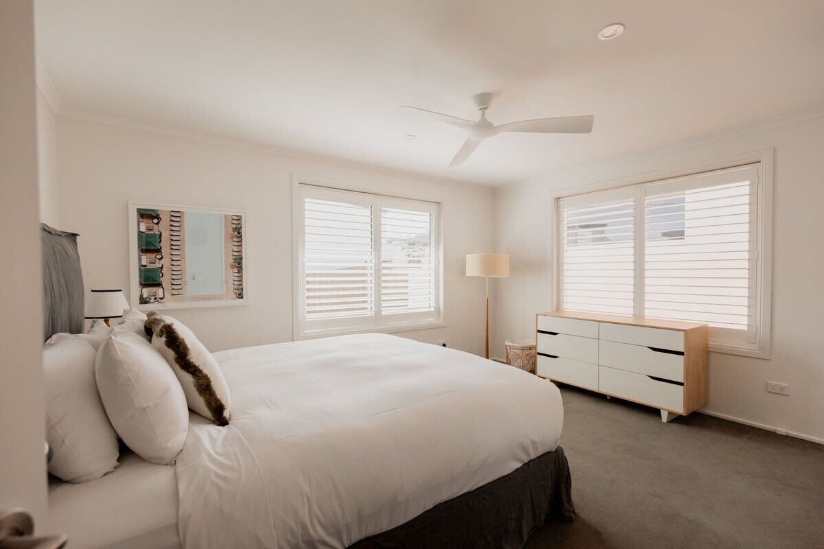 A bedroom with a bed, two windows with blinds, a ceiling fan, a bedside table with a lamp, and a wall-mounted artwork.