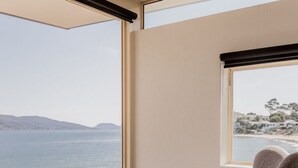Une chambre moderne dotée d’une grande fenêtre donnant sur la côte, et comprenant deux fauteuils gris à revêtement en tissu et un pouf assorti.