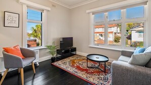 Una sala de estar con televisor, dos sofás, alfombra y ventanas grandes con persianas.