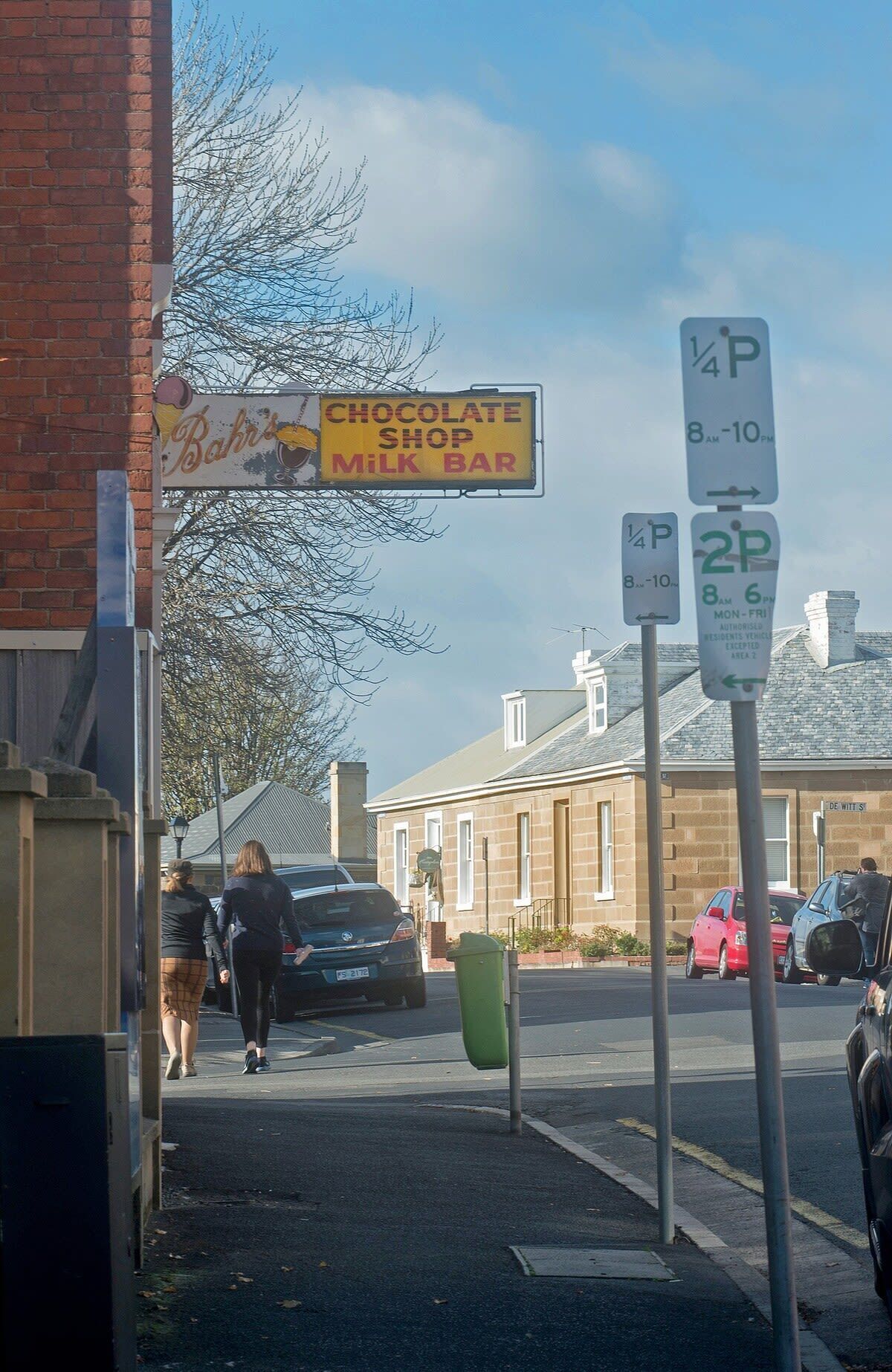 Vista d'un carrer amb un cartell de xocolateria i bar de llet, senyals d'aparcament que indiquen 1/4 i 2/4 d'hora, i una zona residencial amb cases i cotxes aparcats.