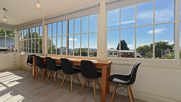 Un espace repas avec une table en bois et des chaises noires, de grandes fenêtres offrant une vue sur les bâtiments et les arbres, et un plancher de couleur claire.
