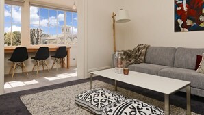 Un salon moderne avec un canapé gris, une table basse blanche et un tapis à motifs. On y trouve trois chaises noires et un bureau avec une lampe. Un tableau est accroché au mur, et une fenêtre offre une vue sur des immeubles et des arbres.