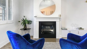 Una sala de estar con chimenea, dos sillones azules, un espejo redondo y una mesita con una planta.