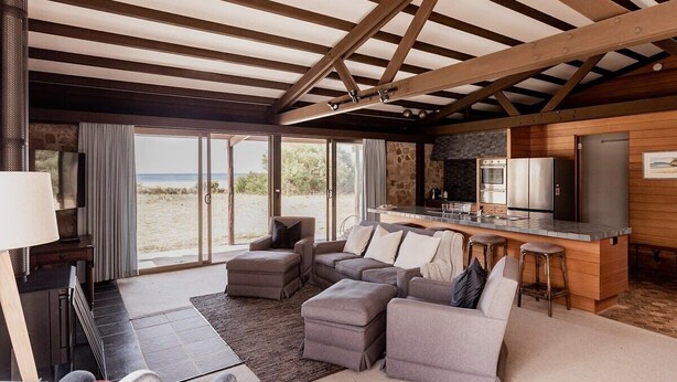 A modern living room with a large sectional sofa, a kitchen area with a bar, and a view of the beach through large sliding doors. - Waterfront Boltons Beach Cottages - room for 8 (Triabunna)