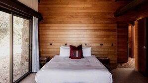 A bedroom with a wooden ceiling, a large bed with white bedding, and a stone wall visible through a glass door. - Waterfront Boltons Beach Cottages - room for 8 (Triabunna)