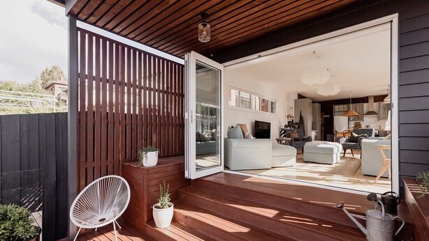 Una terraza de madera con un ventanal corredero que da a una sala de estar con sofá, reposapiés y una mesa de comedor.