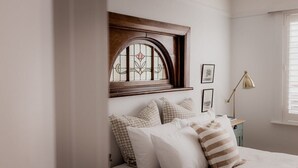Un dormitorio con cama, almohadones estampados, cabecera de madera, una ventana de vitral, un escritorio con lámpara y cuadros en la pared.