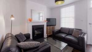 Een moderne woonkamer met een open haard, zwarte leren banken, een glazen salontafel en een televisie op een houten dressoir.