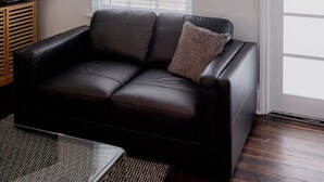 Een woonkamer met een zwarte leren bank, een televisie en een groot raam met zonwering.