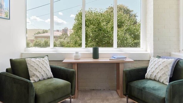 Ein Zimmer mit einem großen Fenster, einem Schreibtisch, zwei grünen Sesseln und einer weißen Ziegelwand.