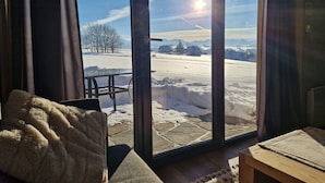 Een kamer met een grote glazen deur die uitkomt op een besneeid gebied, een houten tafel en een gezellige fauteuil met een deken.