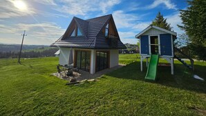 Een huis met een schuin dak, een terras met tuinmeubilair en een speeltuintje met een glijbaan.