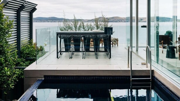 Uma área de piscina moderna ao ar livre, com guarda-corpo de vidro, mesa de jantar com cadeiras e vista para o mar.
