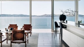 Uma área de refeições moderna com vista panorâmica para o mar, com uma mistura de cadeiras de madeira e couro, uma mesa branca e um grande vaso decorativo.