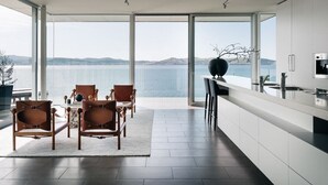 Cozinha moderna com grandes janelas, área de refeições com cadeiras de couro e vista para o mar.
