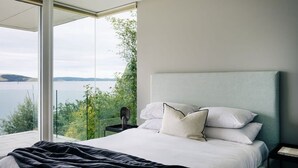 Quarto com uma cama grande, roupa de cama branca e vista para um lago e vegetação.