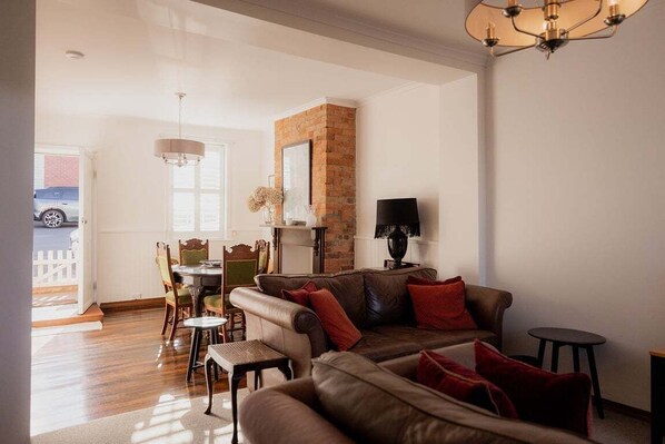 A living room with a brown leather sofa, a dining table with chairs, a brick fireplace, and a chandelier.
