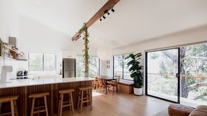 Una cocina moderna con una isla central, taburetes de madera, una zona de comedor con sillas y ventanas amplias que ofrecen vistas al verdor.