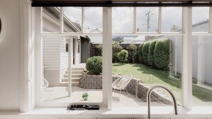A view from a window with a white interior, looking out to a backyard with a lawn, a brick wall, and a white chair.