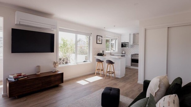 A modern living room with a flat-screen TV, a wooden console table, and a black ottoman. There is a kitchen area with a white island, bar stools, and a large window providing natural light. - Modern refurbished unit in charming Bellerive (Bellerive)
