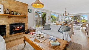A spacious living room with a fireplace, a wooden coffee table, a sofa, and a dining area with natural light from large windows.