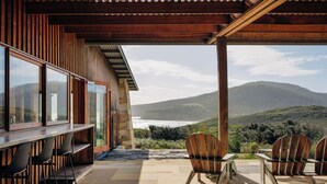 Une terrasse extérieure offrant une vue sur l’océan et les collines, avec des chaises en bois et un espace bar.