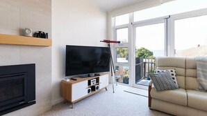 Un salon moderne avec un téléviseur à écran plat posé sur un meuble en bois, une cheminée, un canapé agrémenté d’un coussin à motifs et de grandes fenêtres offrant une vue sur l’extérieur.