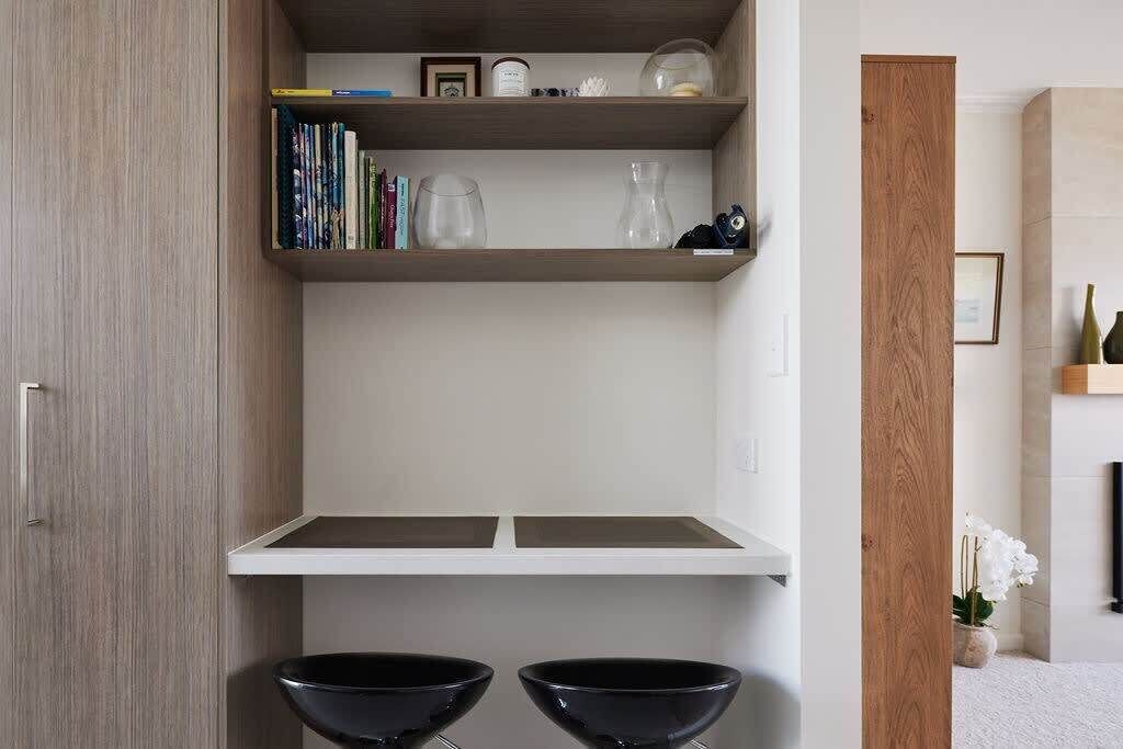 A modern room with a wooden wardrobe, a white shelf with books and decorative items, and two black stools.