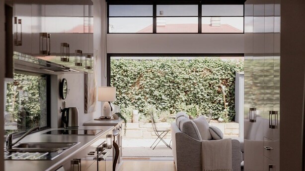 A modern kitchen with a large window, a dining area with a white sofa, and a view of a lush garden. - Stay steps from Salamanca in historical cottage (Battery Point)