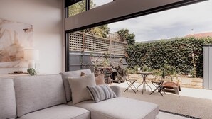 A modern living room with a grey sectional sofa, a small round table, and a chair. There is a large window providing a view of a garden with a lattice fence and greenery. - Stay steps from Salamanca in historical cottage (Battery Point)