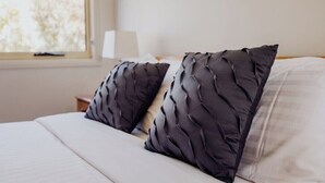 Una cama con dos almohadas oscuras y un colchón blanco, una mesita de noche con una lámpara y una ventana con vistas a árboles.