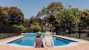 Dos personas sentadas junto a una piscina rectangular con patio de ladrillo, rodeado por una cerca de metal negro y árboles.