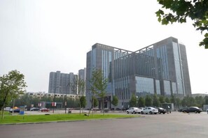 A modern building with a glass facade, surrounded by a parking lot and a few trees. - Guanhe International Hotel (Shijiazhuang)