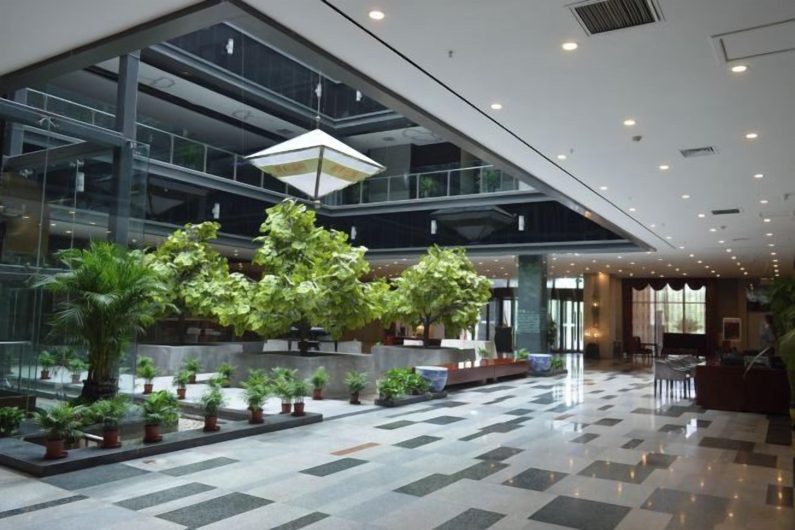 Um hall amplo com design moderno, apresentando uma grande parede de vidro, um arranjo de plantas central e um piso com padrão.