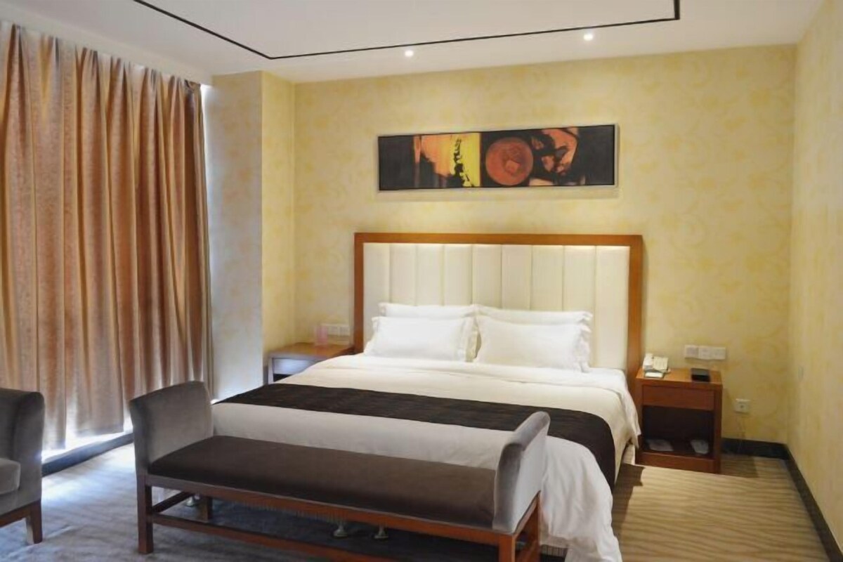Habitación de hotel con cama, mesitas de noche, una silla, un banco, un cuadro en la pared y una cortina.